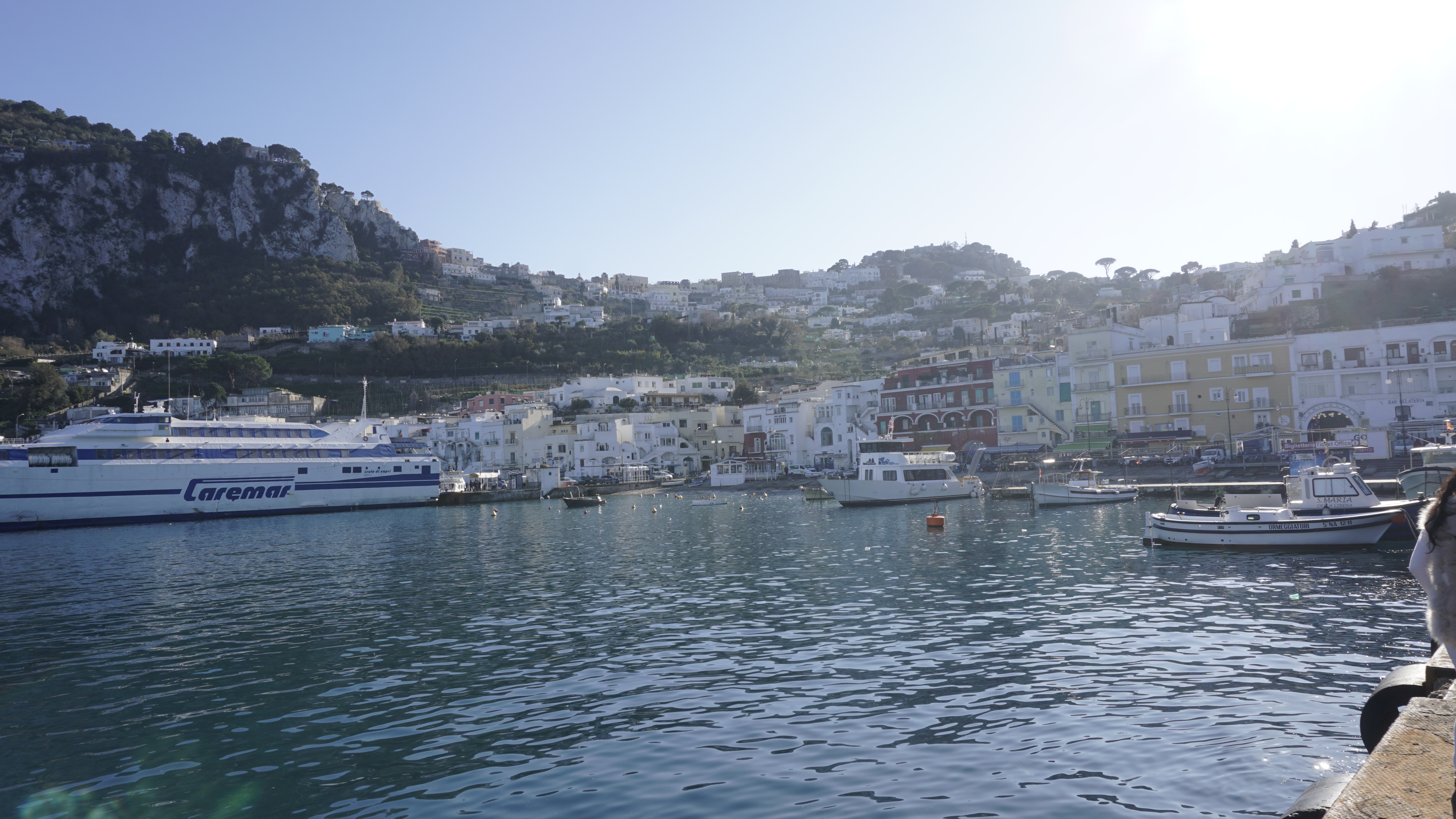 Boats and a ferry in a harbour beneath hillside buildings.
