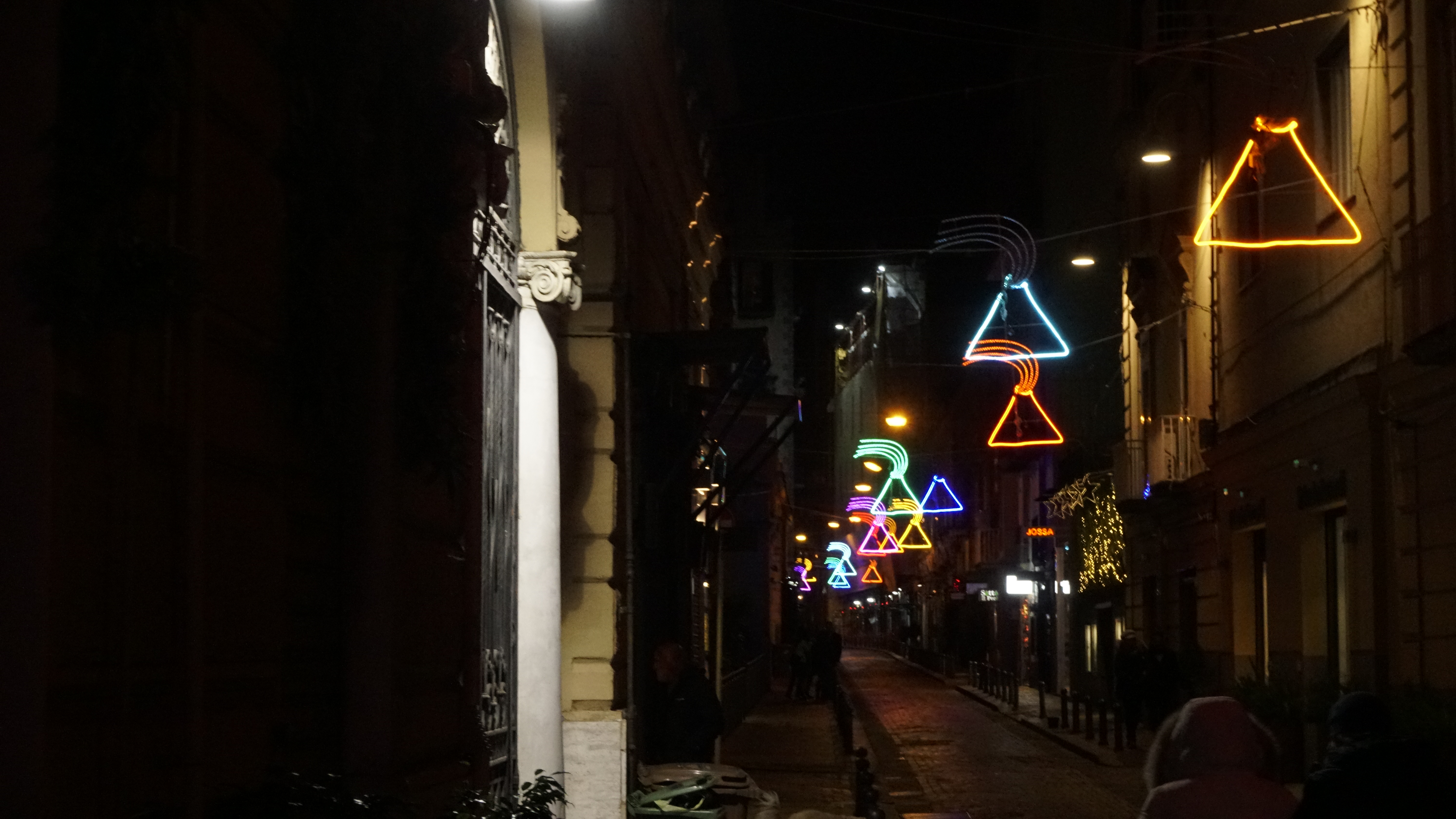 Colourful neon shapes hang above a dark, narrow city street.