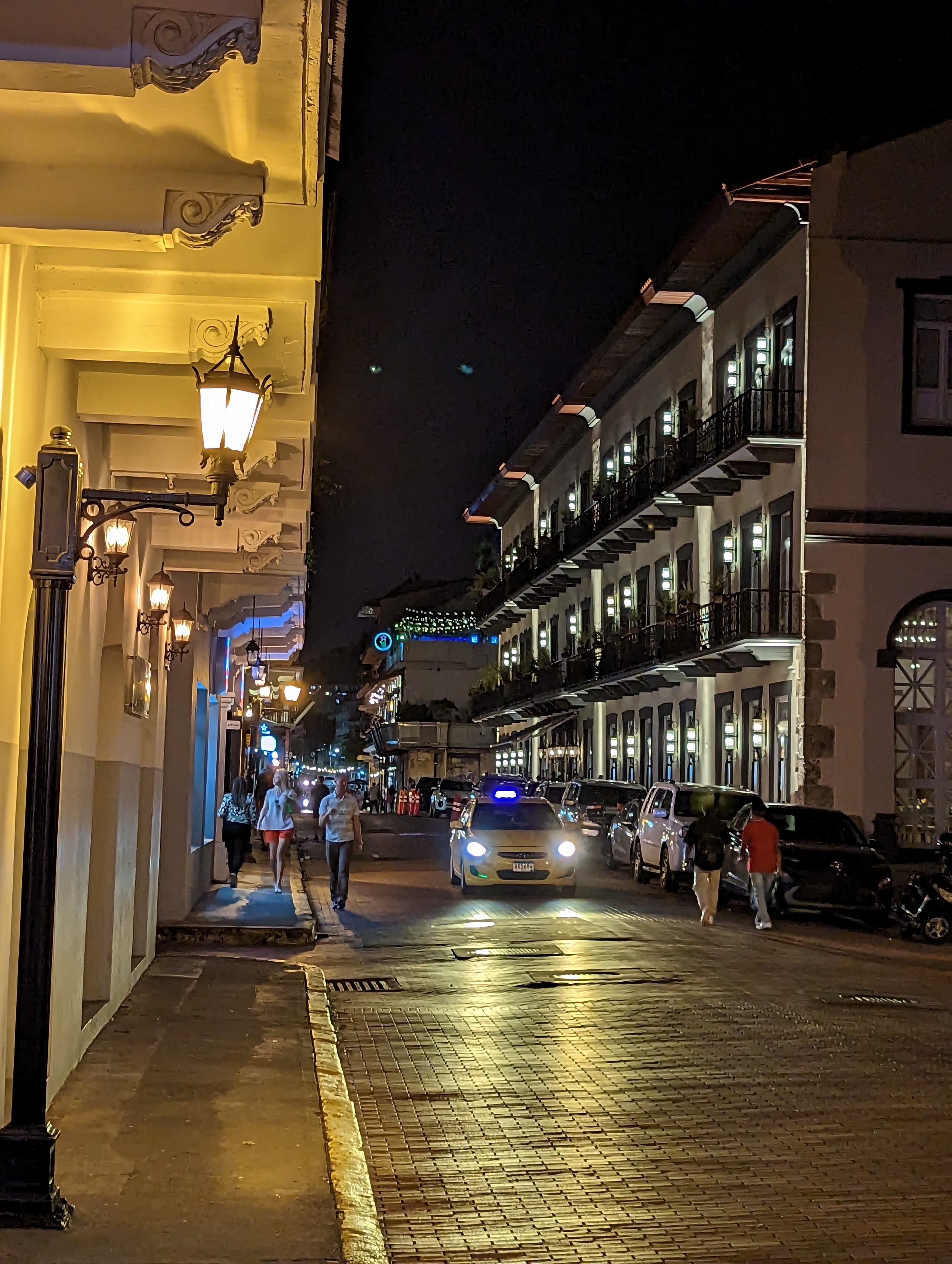 Golden lanterns illuminate a narrow street with balconies and a taxi.